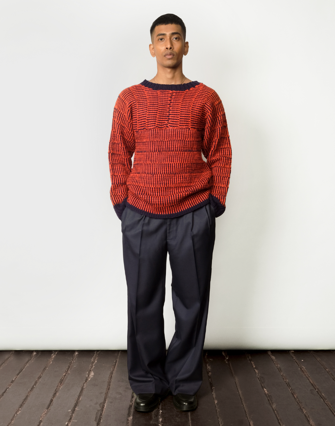 A male model wearing a red and navy striped kimono-cut sweater with a boat neckline and wide leg navy trousers.