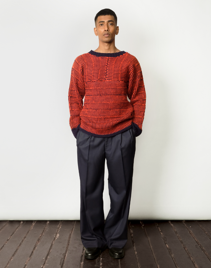 A male model wearing a red and navy striped kimono-cut sweater with a boat neckline and wide leg navy trousers.